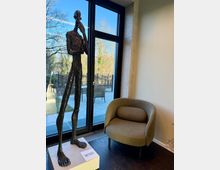 Eine moderne Innenraumszene mit einer schlanken Metallskulptur auf einem Sockel, einem braunen Sessel und einer großen Glasfensterfront mit Blick auf einen Gartenbereich. Durch die Fenster sind Bäume und eine geflieste Terrasse mit Stühlen zu sehen.