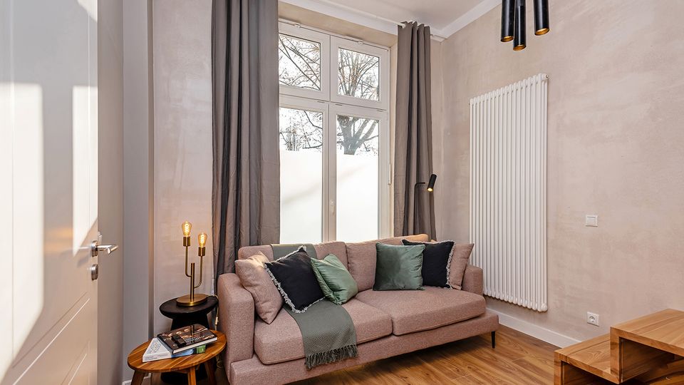 Modernes Wohnzimmer mit hellen Wänden, Holzboden und einem Sofa vor hohen Fenstern mit grauen Vorhängen. An der Wand hängt ein großer weißer Heizkörper; daneben stehen ein kleiner Beistelltisch mit Lampe und ein niedriger Couchtisch, darüber schwarze Pendelleuchten.