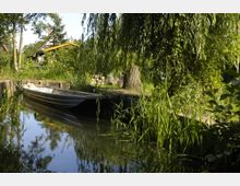 Ein ruhiger Wasserkanal mit einem schmalen Holzboot am Ufer, umgeben von dichter Vegetation wie Weidenbäumen und hohem Gras; im Hintergrund ist ein Haus mit gelbem Dach zu sehen.