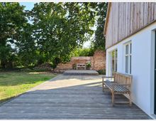 Holzterrasse neben einem modernen Gebäude mit weißer Fassade und großen Fenstern, im Hintergrund eine rote Backsteinmauer mit dekorativen Pflanzen davor; umgeben von Bäumen und einer Rasenfläche. Eine Holzbank steht an der Gebäudewand.