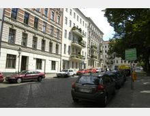 Straßenszene in einem Wohnviertel mit mehrstöckigen Altbauten, teilweise mit Balkonen und Blumen. Autos parken entlang der Kopfsteinpflasterstraße, und ein Baumreihe spendet Schatten; ein Schild weist auf einen Wochenmarkt hin.