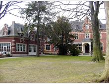 Große Backsteinvilla mit historischen Fassadendetails und hohen Fenstern, umgeben von Bäumen und einer Rasenfläche. Das Gebäude hat ein steiles Schieferdach und einen zentralen Eingang mit einer überdachten Veranda.