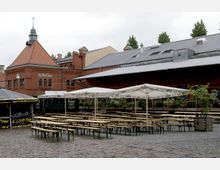 Ein Platz mit Kopfsteinpflaster, gesäumt von Bierbänken und Tischen unter weißen Sonnenschirmen. Im Hintergrund stehen historische Backsteingebäude mit roten Ziegeldächern und Schornstein, darunter ein Gebäude mit der Aufschrift „Pool & Cigars“.