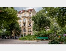 Eine historische Wohnhausfassade mit gelbem Putz und dekorativen Balkonen steht hinter einer kleinen Parkfläche mit Bäumen, Sträuchern und einer grünen Parkbank. Im Vordergrund führen Gehwege durch die grüne Umgebung, und ein Straßenschild zeigt den Namen „Suarezstraße“.