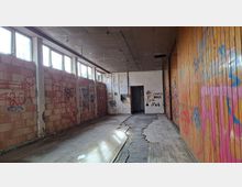 Langer, leerer Innenraum einer verlassenen Industriehalle mit hohen Fensterreihen links, rauen Ziegelwänden und einer holzverkleideten Wand rechts, die mit Graffiti besprüht ist. Der Boden ist teilweise beschädigt und aufgerissen, am Ende des Raums befindet sich eine offene Tür in einer weißen Wand.