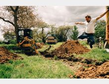 Eine Baustelle auf einer Wiese mit einem kleinen gelben Bagger, der in einem Graben arbeitet. Im Hintergrund sind Bäume und ein alter Anhänger zu sehen, während rechts im Bild eine Person in die Luft springt.