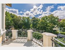 Balkon eines Wohnhauses mit hellen Pfeilern und dekorativem schmiedeeisernem Geländer, mit Blick auf eine von hohen Bäumen gesäumte Wohnstraße. Im Hintergrund sind weitere Häuserdächer unter blauem Himmel mit Wolken zu sehen.