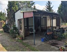 Kleines eingeschossiges Gartenhaus mit Flachdach und gemischter Fassade aus Holz, Stein und großen Fensterflächen. Davor befindet sich eine überdachte Terrasse mit Metallstützen, Sitzmöbeln und Topfpflanzen, umgeben von Bäumen und einem einfachen Gartenzaun.