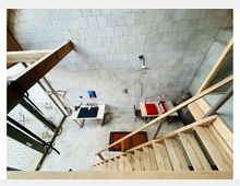 Innenansicht eines hohen, minimalistischen Raums mit hellen, kleinteiligen Wandfliesen, Sichtbetonboden und einer Holztreppe mit Geländer. Große bodentiefe Fenster lassen Tageslicht herein; darunter stehen ein schmaler Tisch, ein Stuhl und wenige weitere Möbel.