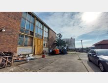 Industriegebäude mit einer Fassade aus Ziegelstein und großen, vergitterten Fenstern, daneben ein Hof mit abgestellten Müllcontainern, Holzstapeln und Baumaterialien. Im Hintergrund sind ein Wohngebäude, ein Zaun und geparkte Autos zu sehen, bei sonnigem Wetter mit leicht bewölktem Himmel.