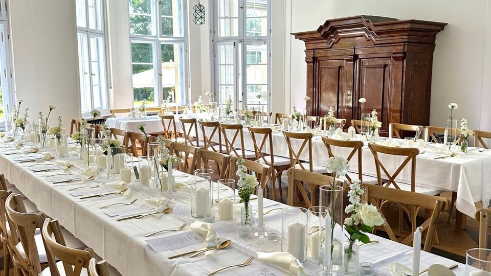 Heller Festsaal mit hohen, bodentiefen Sprossenfenstern und dekorativem Stuck an der Decke, ausgestattet mit langen, weiß gedeckten Tafeln und Holzstühlen. An einer Wand steht ein großer, dunkler Holzschrank; der Raum hat einen Holzboden und schlichte weiße Wandpaneele.