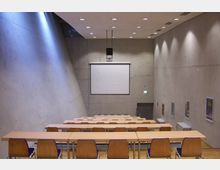 Kleiner Hörsaal mit einer Betonwand-Architektur, einer Projektionsleinwand an der Stirnseite und stufenförmig angeordneten Holztischen und Stühlen. Der Raum ist minimalistisch gestaltet, mit schrägen Wänden und mehreren Wandplakaten.