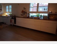 Helle Küche mit weißen Unterschränken und einer dunklen Holz-Arbeitsfläche, auf der Geschirr, Obst und Pflanzen stehen. Zwei Fenster bieten einen Ausblick auf Häuser mit roten Dächern und Außengrünflächen.