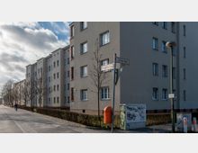 Straßenansicht eines Wohnviertels mit mehrstöckigen, hellgrauen Wohnhäusern im schlichten Stil. Am Gehsteig stehen Bäume ohne Laub, eine Straßenlaterne, ein orangefarbener Briefkasten und ein Schaltkasten mit Graffiti, daneben Straßenschilder mit den Namen "Zeiler Weg" und "Planbauern Promenade".