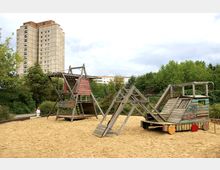 Spielplatz mit hölzernen Klettergerüsten und einem sandbedeckten Boden, umgeben von Bäumen und Büschen; im Hintergrund ein mehrstöckiges Hochhaus und weitere Wohngebäude.