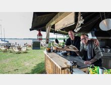 Eine offene Strandbar aus Holz mit Blick auf einen See. Im Vordergrund befindet sich eine Theke mit Kochutensilien, im Hintergrund sind Liegestühle, Sonnenschirme und Boote am Ufer zu sehen.