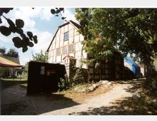 Fachwerkhaus mit hellem Putz und dunklen Holzbalken, umgeben von Bäumen und Garten. Auf der linken Seite ein kleiner schwarzer Schaukasten, rechts ein geschwungener Kiesweg.