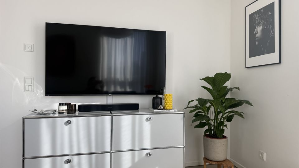 Moderne Wohnzimmerecke mit großem Flachbildfernseher auf einem silbernen Sideboard, darauf Soundbar und kleine Dekoobjekte. Rechts steht eine Zimmerpflanze im Topf neben einem gerahmten Schwarzweißfoto an der weißen Wand.