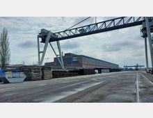 Industriegelände mit einem großen Lagergebäude aus Backstein und Glas, auf dem das Schild „Mebra Remine“ zu sehen ist. Im Vordergrund befindet sich ein Kran auf Schienen sowie gestapelte Materialien, im Hintergrund weitere industrielle Anlagen und Geräte.