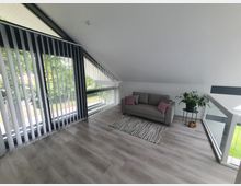 Heller Wohnbereich unter einer Dachschräge mit großen, bodentiefen Fenstern und vertikalen Lamellenjalousien. Der Raum verfügt über einen grauen Sofa, Teppichboden, eine Zimmerpflanze auf einem kleinen Tisch und eine Glasabtrennung, die einen Blick nach unten gewährt.