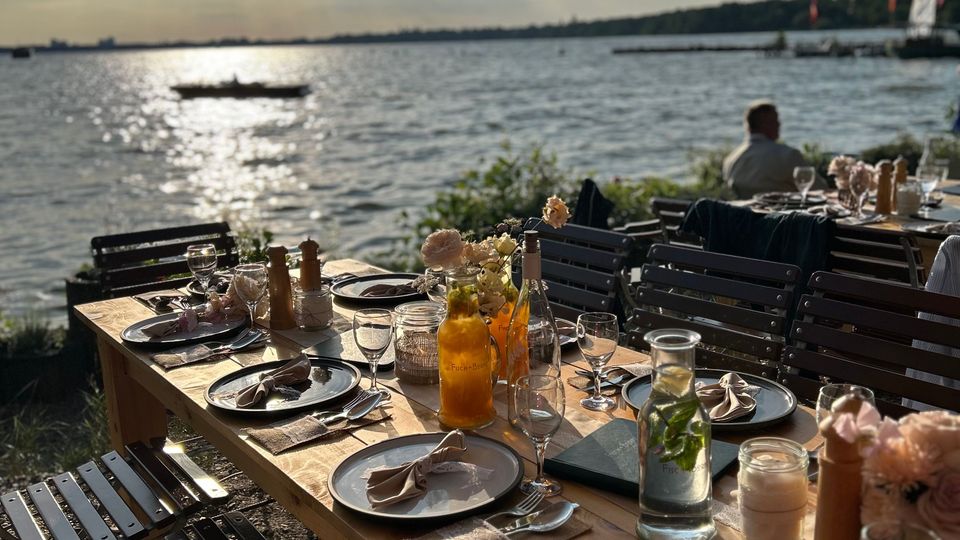 Großer, gedeckter Holztisch im Freien mit eleganter Tischdekoration, darunter Blumen, Stoffservietten und Glasflaschen, am Ufer eines Sees mit Blick auf das Wasser, unter einem sonnigen Himmel mit Wolken.