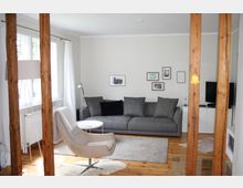 Wohnzimmer mit hellen Wänden und Holzfußboden, eingerichtet mit einem grauen Sofa, einem hellgrauen Sessel und einem kleinen runden Couchtisch auf einem Teppich. An der Wand hängen mehrere kleine Bilder, und die Zimmertrennung erfolgt durch vertikale Holzbalken.