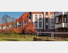 Historisches Backsteingebäude mit weißen Verzierungen und roten Ziegeldächern, umgeben von einer gepflegten Gartenanlage mit einem kleinen Baum voller roter Beeren im Vordergrund. Im Hintergrund sind kahle Bäume und weitere Gebäude zu sehen.