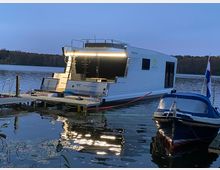 Ein modernes Hausboot mit beleuchteter Terrasse liegt an einem Steg auf einem ruhigen See, daneben ist ein kleines Motorboot festgemacht. Im Hintergrund ist eine bewaldete Uferlandschaft zu sehen, leicht von der Dämmerung beleuchtet.