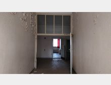 Ein leerer, leicht heruntergekommener Flur mit abblätternder Farbe an den Wänden, der zu einem Raum mit einem roten Vorhang und Tageslicht führt. Über der Tür befindet sich eine Glaspartie mit kleinen rechteckigen Fenstern, und am Boden liegen Kabel verstreut.