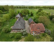 Luftaufnahme eines ländlichen Hofensembles mit reetgedeckten Fachwerkgebäuden und einem roten Ziegeldachhaus, umgeben von Bäumen, Wiesen und Feldwegen. Im Hintergrund erstrecken sich offene Felder bis zum Horizont unter bewölktem Himmel.