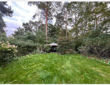 Grüne Gartenlandschaft mit einer kleinen Wiese im Vordergrund, umgeben von Bäumen und Sträuchern; in der Mitte befindet sich ein Pavillon mit metallischem Dach, eingebettet in das dichte Pflanzenwerk.