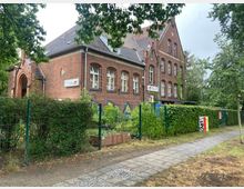 Backsteingebäude mit spitzem Giebeldach, umgeben von einem Zaun und dichter Begrünung. Im Eingangsbereich ein kleiner Torbogen, davor Gehweg und teilweise Rasenflächen; Schilder an der Fassade weisen auf eine Kindereinrichtung hin.