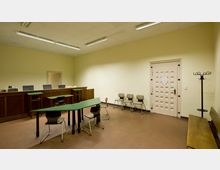 Ein kleiner Raum mit beigefarbenen Wänden und einfacher Möblierung, darunter Tische mit grünen Oberflächen, Stühle, eine Holzbank und ein Holztresen. Eine helle Tür mit quadratischen Aussparungen und Hakenständer an der Seite ergänzen die Ausstattung.