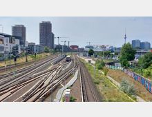 Bahngelände mit mehreren Gleisen und einer vorbeifahrenden S-Bahn, umgeben von moderner Stadtbebauung. Im Hintergrund sind Hochhäuser, Baukräne, das Mercedes-Benz-Arena-Gebäude und der Berliner Fernsehturm zu sehen.