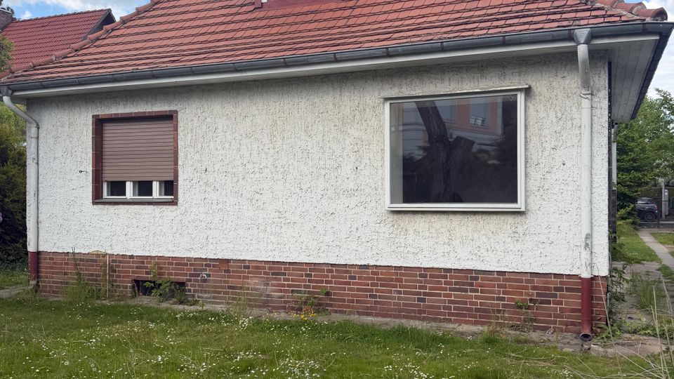 Seitenansicht eines eingeschossigen Wohnhauses mit weiß verputzter Fassade, rotem Ziegeldach und kleinem Dachgaubenfenster. Vor dem Haus liegt eine Rasenfläche; sichtbar sind zwei Fenster, eine Regenrinne und ein Sockel aus rotem Backstein.