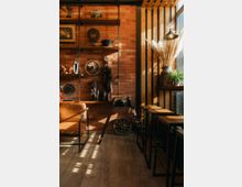 Eine gemütliche Innenraumecke mit Backsteinwand, Holzregalen und rustikaler Dekoration, darunter Metallräder und getrocknete Pflanzen. Links steht ein lederner Sessel, rechts eine Reihe von hohen Holzhockern an einem Tresen, beleuchtet durch einseitig einfallendes Tageslicht.