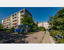 Mehrstöckige Wohngebäude aus Beton mit Balkonen, umgeben von Bäumen und Sträuchern. Im Vordergrund sind ein Gehweg, Sitzbänke sowie ein umzäunter Bereich mit Müllcontainern zu sehen.