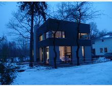 Modernes zweistöckiges Wohnhaus mit dunkelgrauer Fassade und großen Fenstern, beleuchtet von Innenlicht; umgeben von schneebedecktem Boden, kahlen Bäumen und einer winterlichen Abenddämmerung.