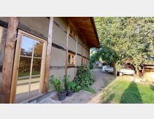 Ein Fachwerkhaus mit Holz- und Lehmelementen, großen Fenstern und einem kleinen bepflanzten Bereich davor, umgeben von einem Hof mit Bäumen, Grünflächen und geparkten Anhängern im Hintergrund.