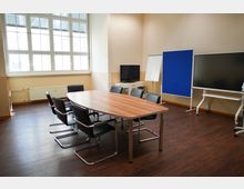 Konferenzraum mit einem ovalen Holztisch, umgeben von schwarzen Bürostühlen. Im Hintergrund befinden sich ein großes Fenster, ein Flachbildfernseher, ein Flipchart, eine blaue Pinnwand und ein großer Monitor auf einem Rollgestell.