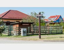 Ein kleines Gehege mit einem Schild "Entenhausen", umgeben von einem Zaun und flankiert von Wohnhäusern mit roten und blauen Dächern im Hintergrund; im Vordergrund ein Wegweiser mit den Straßennamen "Eulenweg" und "Mühlenweg".