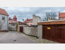 Gepflasterter Gehweg zwischen Wohnhäusern und Garagen auf eng bebauten Innenhofflächen; im Hintergrund sind Gebäude mit roten Ziegeldächern sowie der Turm einer Kirche sichtbar.