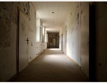 Ein leerer, verlassener Flur in einem heruntergekommenen Gebäude mit abblätternder Farbe an den Wänden. Der Gang ist gerade, mit geschlossenen Türen auf beiden Seiten und schwach beleuchtet durch kleine Fenster.