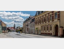 Straßenansicht einer Kleinstadt mit historischen Wohnhäusern, die verputzte und rot verklinkerte Fassaden sowie verzierte Fensterelemente aufweisen; im Hintergrund erhebt sich der Turm einer Kirche unter einem bewölkten Himmel.
