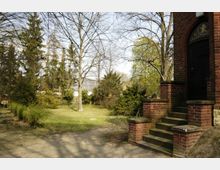 Rasenfläche mit Bäumen und Büschen in einem Garten, rechts im Bild eine Ziegeltreppe mit dunkler Holztür und Rundbogen, kombiniert mit einem dekorativen Fenster. Im Hintergrund sind weitere Gebäude und hohe Bäume zu erkennen.
