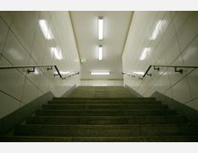 Treppenaufgang in einem U-Bahnhof mit hellen, glänzenden Wandfliesen, Metallgeländern und klarer Deckenbeleuchtung. Ein grünes Notausgangsschild ist an der oberen Wand sichtbar.