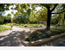 Ein gepflegter Park mit gepflasterten Wegen, grünen Rasenflächen, Bäumen und Blumenbeeten. Im Hintergrund ist eine orange-weiße skulpturale Spitze sowie ein rotes Gebäude sichtbar, während im Vordergrund ein Metallzaun die Szene begrenzt.