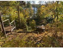 Waldlandschaft im Herbst mit buntem Laub, einem kleinen Holzunterstand und einem Geländer aus Holz. Im Hintergrund ist ein See durch die Bäume hindurch sichtbar.