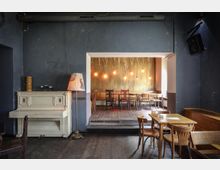 Ein gemütliches Café mit dunklen Wänden und Holzboden; links ein weißes Klavier mit einer Stehlampe daneben, rechts ein erhöhtes Podest mit Holztischen und Stühlen vor einer goldfarbenen Wand mit warmen, hängenden Glühbirnen.