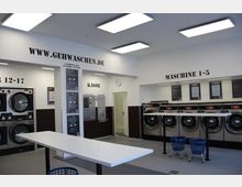 Innenraum eines modernen Selbstbedienungs-Waschsalons mit großen Waschmaschinen und Trocknern. An der Wand befinden sich Beschilderungen, unter anderem zur Anleitung und Anleitungskasse, mit einem zentralen Tisch und Wäschekörben auf Rollen im Vordergrund.